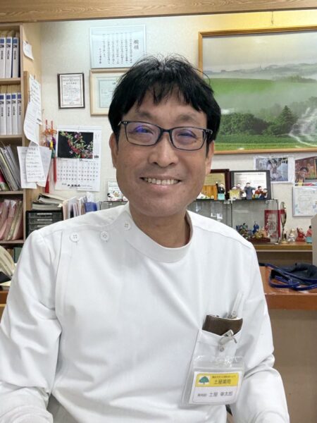 山形県東根市の土屋薬局で、薬剤師の土屋幸太郎さんが笑顔で応対している様子。白衣を着てデスクに座り、温かい雰囲気の店内には季節の風景画や薬棚が見える。信頼感と親しみやすさを感じる薬局の一コマ。