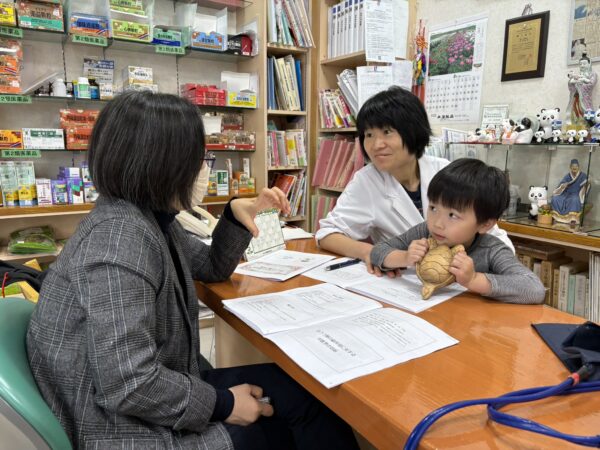 楊 紅娜先生の話を土屋薬局店内で勉強する妻の幸先生と息子