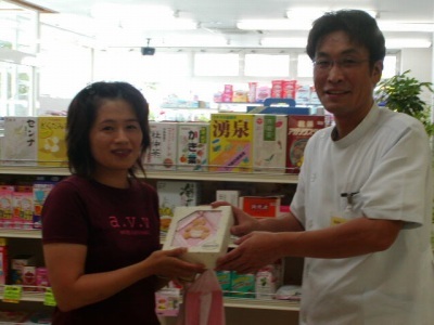 出産後のお客様に、薬剤師が笑顔でピンクのアルバムを手渡している様子。高プロラクチン血症の漢方周期療法で妊娠・出産された方へのお祝いの場面。実際の写真です。
