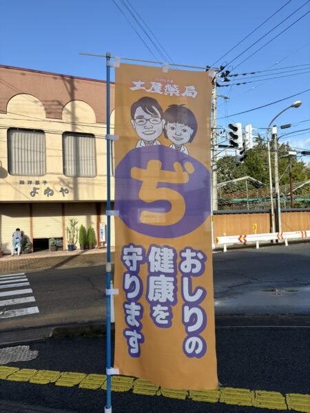 山形県東根市の土屋薬局前に掲げられたのぼり旗。薬剤師夫婦のイラストの下に「ぢ おしりの健康を守ります」と書かれ、痔の漢方相談を行う薬局であることを示しています。