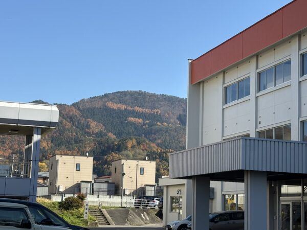 リンクむらやまの敷地越しに、11月の紅葉で色づいた山々が広がる秋の風景。晴れた一日に映える美しい山形の自然景観を写した写真。