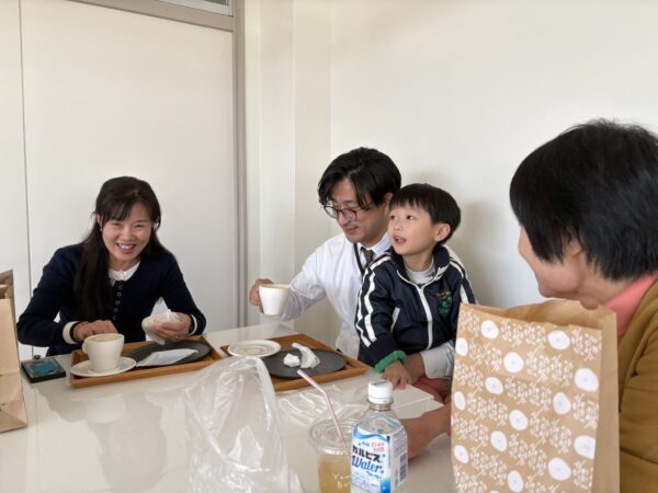 妊活講演会が始まる前、講師やスタッフ、家族、担当者が和やかにベーグルを食べながら笑顔で談笑している様子。リラックスした雰囲気が伝わる準備中の風景。