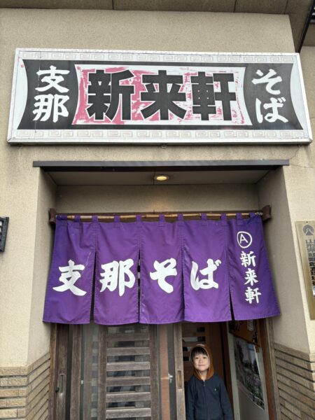 長井市の新来軒ののれんの下で、4歳の息子がにっこり笑顔で立っている写真。お店の前で嬉しそうにポーズをとる姿が微笑ましい一枚。