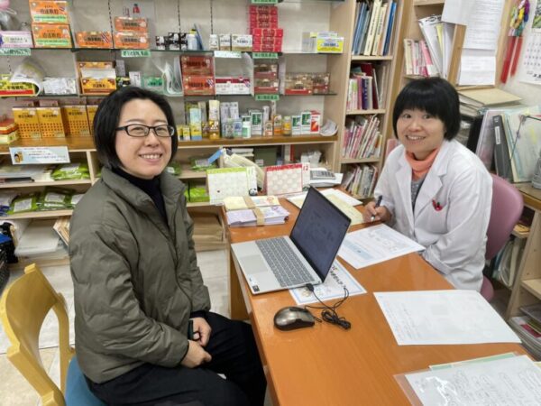 土屋薬局の店内で、漢方の専門家・楊紅娜（ようこうな）先生と土屋幸先生が学び合っている様子。亀鹿仙や冠元顆粒など女性の体質改善に役立つ漢方について意見を交わしている。明るく信頼感のある店内の雰囲気が伝わる写真。