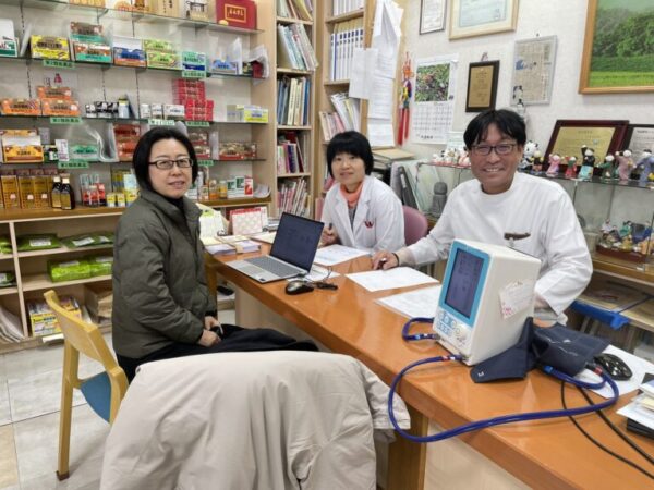 土屋薬局の店内で、薬剤師の土屋幸太郎、妻の土屋幸先生、中医師の楊紅娜（ようこうな）先生がにっこり笑顔で並んでいる写真。亀鹿仙や冠元顆粒などの漢方を通じて学びを深め合う、温かく信頼感のある雰囲気が伝わる一枚。