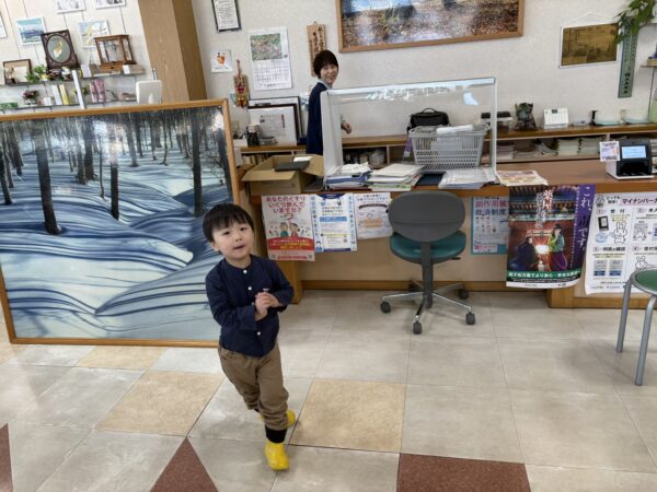 土屋薬局店内で、３歳の息子が黄色い長靴をはいて冬の絵の前で微笑む写真。令和６年１２月１０日撮影。山形の冬らしい温かい雰囲気の店内風景。