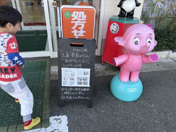 土屋薬局の店頭に設置された案内看板の前で、サトコちゃん人形と一緒に元気よく立っている年少さんの息子の写真。明るい表情で手を振る姿が、家族経営の温かさと welcoming な雰囲気を伝えている。