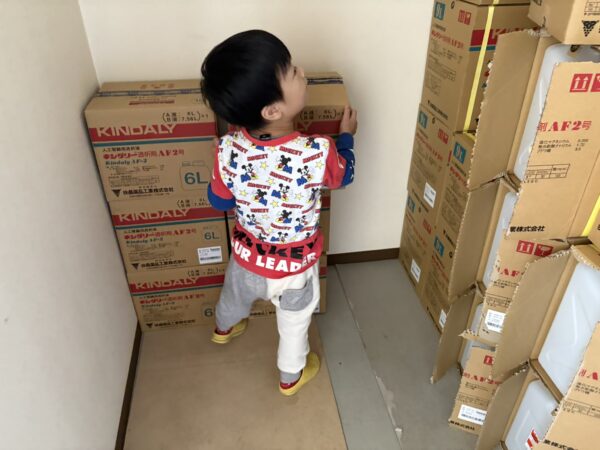 土屋薬局の倉庫で、4歳の息子が透析液の空き箱を両手で運んでいる様子。小さな体で一生懸命お手伝いをする姿が、家族経営の温かさと優しい雰囲気を伝えている写真。