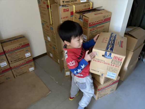 透析液の空き箱を抱えて歩く4歳の息子の後ろ姿を写した写真。倉庫の棚の間を一生懸命に運ぶ姿が、薬局の日常や子どもの成長を感じさせる温かい場面。