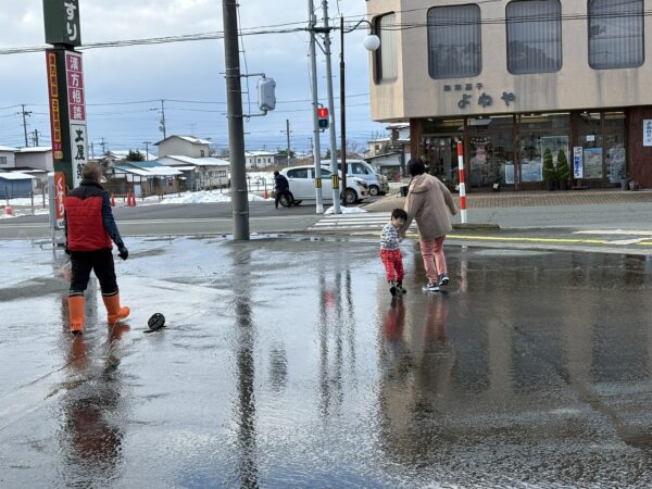 駐車場の融雪用の井戸水が流れるか家族で点検している様子。寒い中、妻と息子が手を赤くしながら作業を手伝う心温まる冬のシーン。