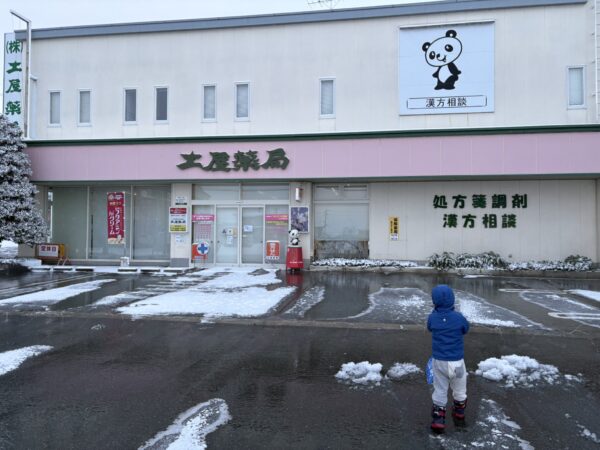 冬の朝、山形県東根市の土屋薬局の駐車場で、子どもが背中を向けて雪かきをしている様子
