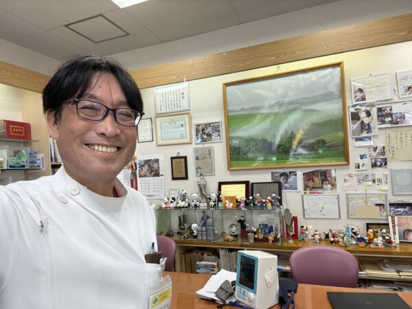 山形県東根市の土屋薬局の子宝相談コーナー。妊活相談を行う薬剤師・土屋幸太郎と、壁面に並ぶ出産報告の赤ちゃんの写真。