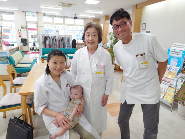 山形県東根市の土屋薬局にて、白衣姿で笑顔を見せる薬剤師の土屋幸太郎さんとご家族。赤ちゃんを囲みながら、地域に寄り添った漢方相談や妊活相談を行っている様子。