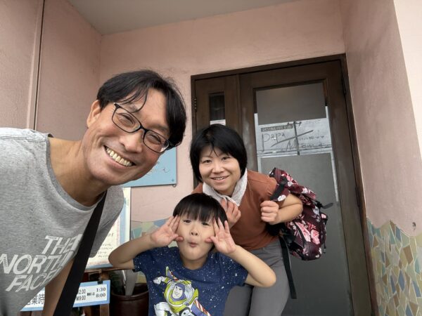 2026年7月13日、東根市蟹沢の洋食店「パパアンドママ」の入口で記念撮影をする土屋薬局の家族3人。ランチを楽しんだ後の笑顔の一枚。