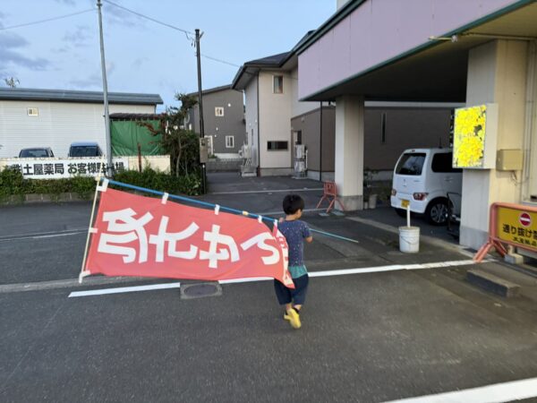 土屋薬局の店先で、運動会を終えた5歳の息子がのぼりの片付けを手伝っている様子
